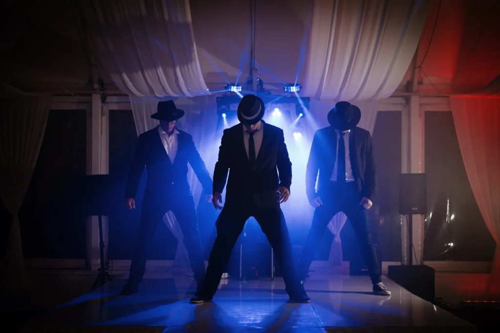 Three performers from the Men of Steel dressed in suits and hats, standing on stage with dramatic lighting at Allely Estate.