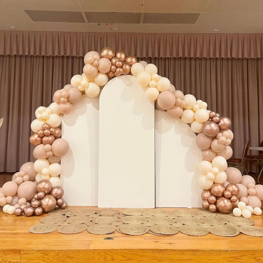 Organic balloon decor in neutral blush, beige, and rose gold metallic tones over three white arched backdrop plinths, styled for a modern Auckland event.