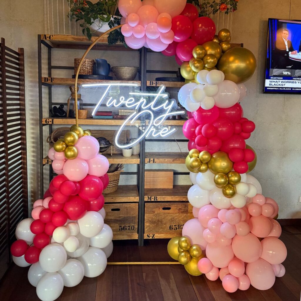 Organic balloon decor installation in pink, red, and gold, featuring a neon "Twenty One" sign. The arch is styled for a 21st birthday party in Auckland.