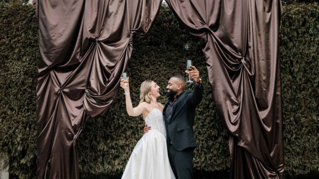 Elegant wedding couple celebrating with champagne under dramatic luxury drapery captured by HAP FILM with cinematic wedding photography and videography.